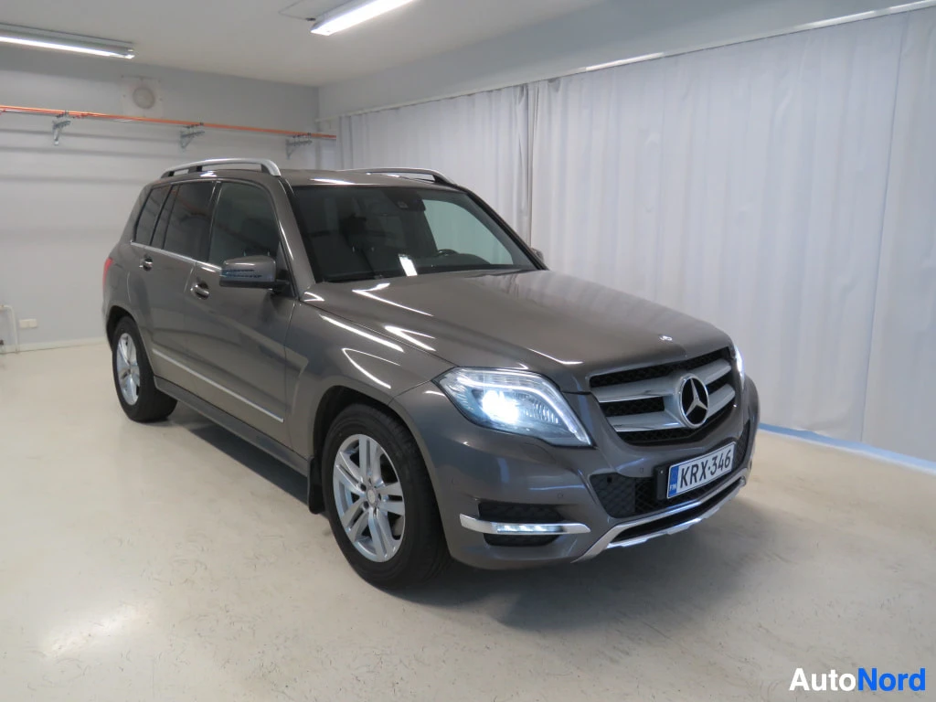 Etuviisto, nokka oikealle: Mercedes-Benz GLK:n etukulmanosto: keulagrilli merkillä, ajovalot, kylki ja rekisterikilpi näkyvät valkoista verhoseinää vasten sisätiloissa.