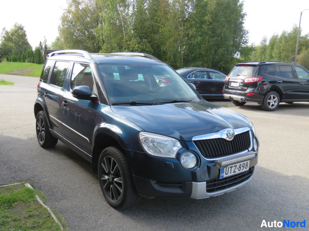 Etuviisto, nokka oikealle: Tummansininen Škoda Yeti edestä vasemmalta kolmannelta kuvattuna, mustat vanteet ja kattokaiteet.