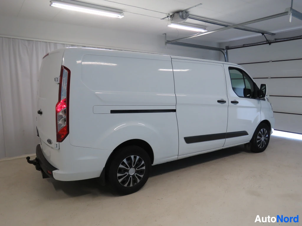 Takaviisto: Valkoinen Ford Transit Custom -pakettiauto takaa ja vasemmalta sivulta kuvattuna garmaasissa, takana vetokoukku.