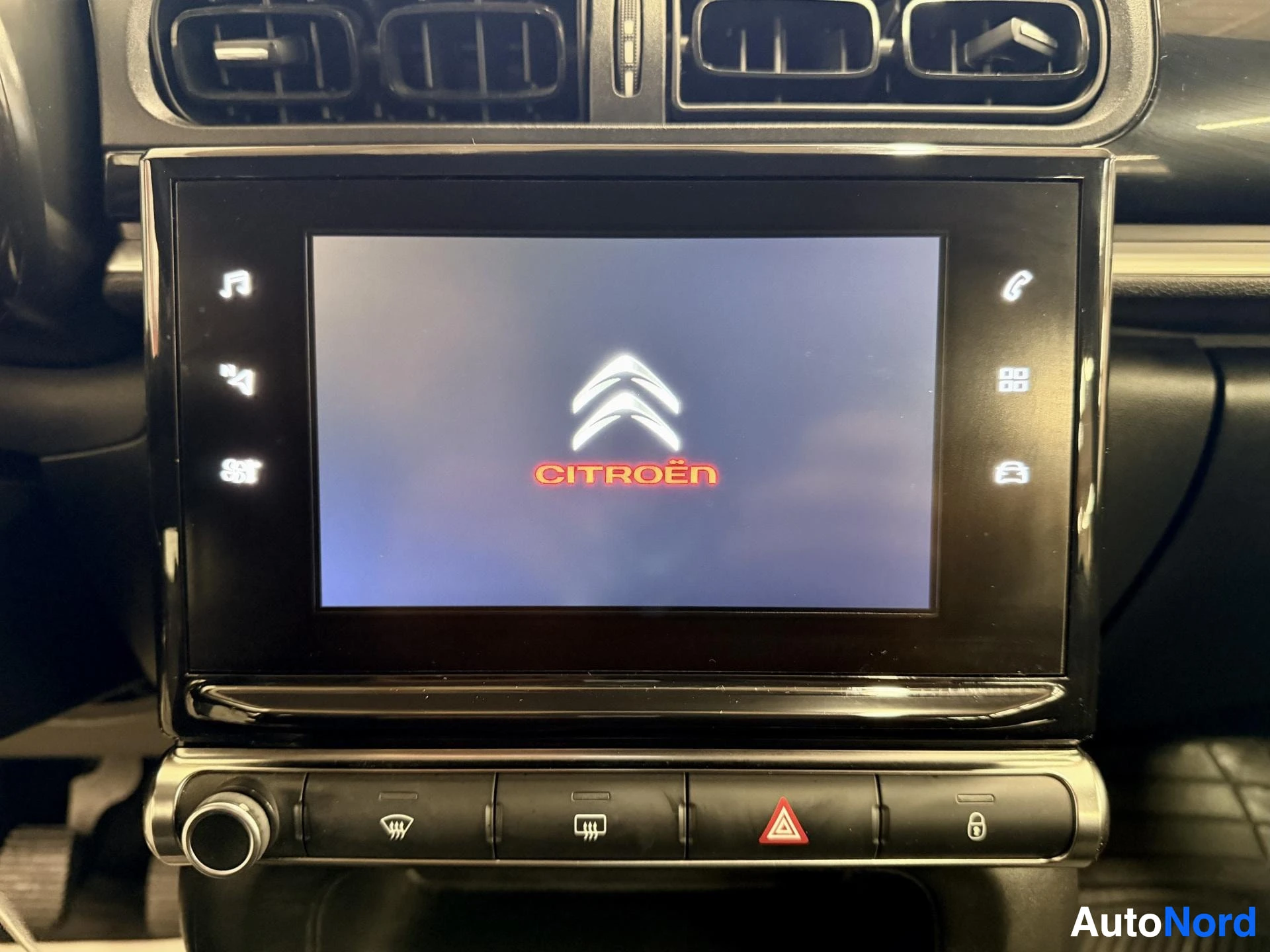 Yksityiskohta: Citroënin kojelaudan kosketusnäyttö käynnistymässä, ruudussa Citroën-logo; alapuolella paneeli pienillä painikkeilla.