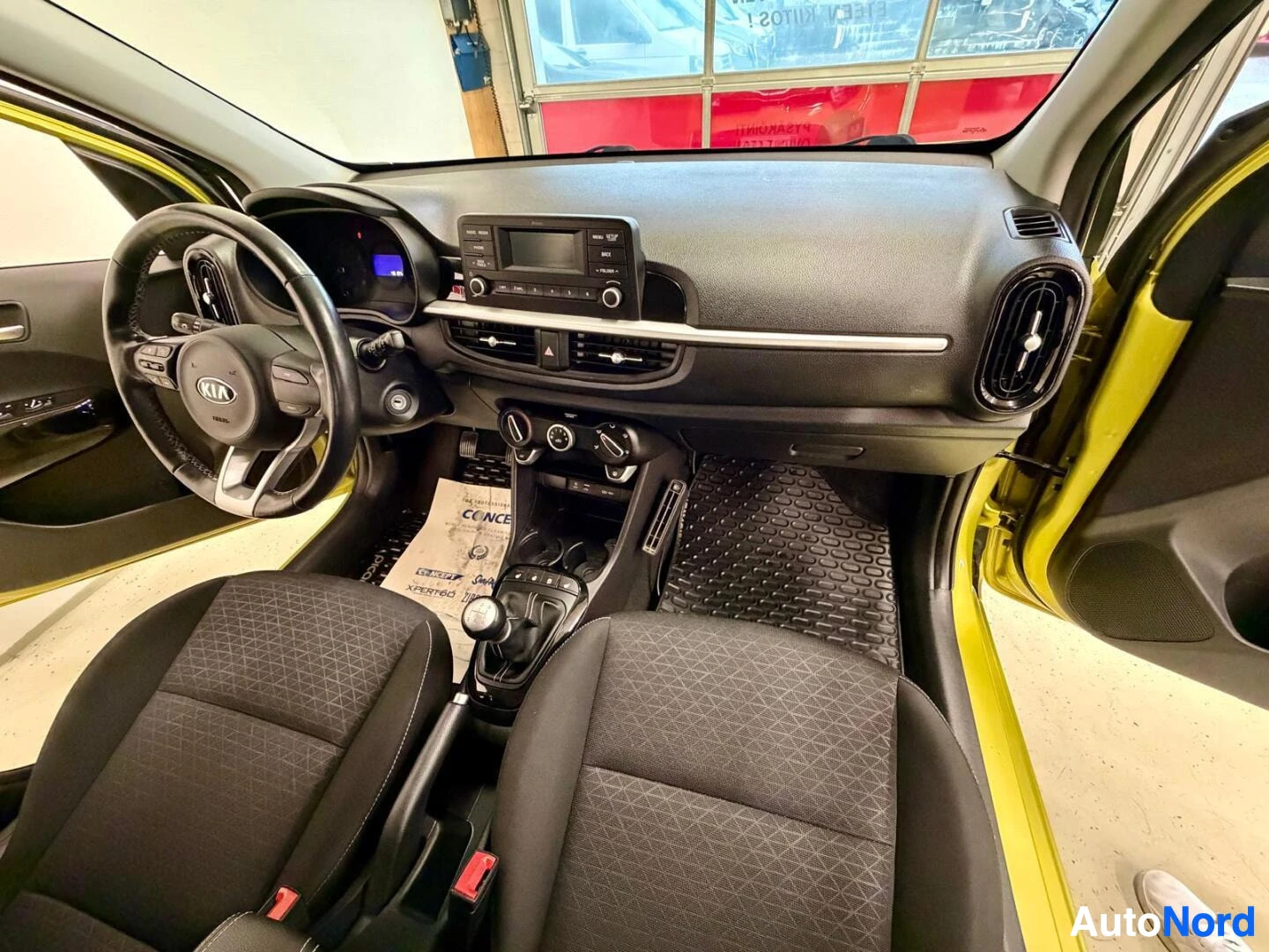 Edestä: Auto kuvattuna sisältä: musta kojelauta, Kia-merkitty ohjauspyörä, radio, manuaalivaihtokeppi ja kaksi tummaa kangasetuistuinta; kumimatot lattiassa ja ovista p