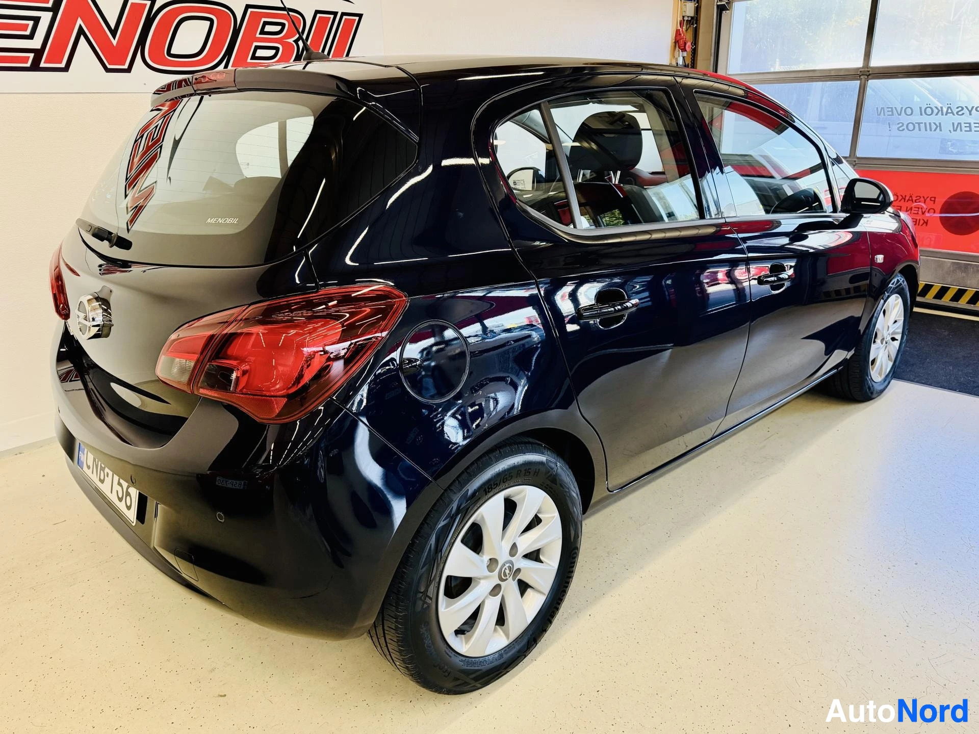 Takaviisto: Mustan sinertävän tumma Opel Corsa -henkilöauto takaviistosta kuvattuna liiketilan lattialla; takavalo, takapuskuri ja rekisterikilpi näkyvät.