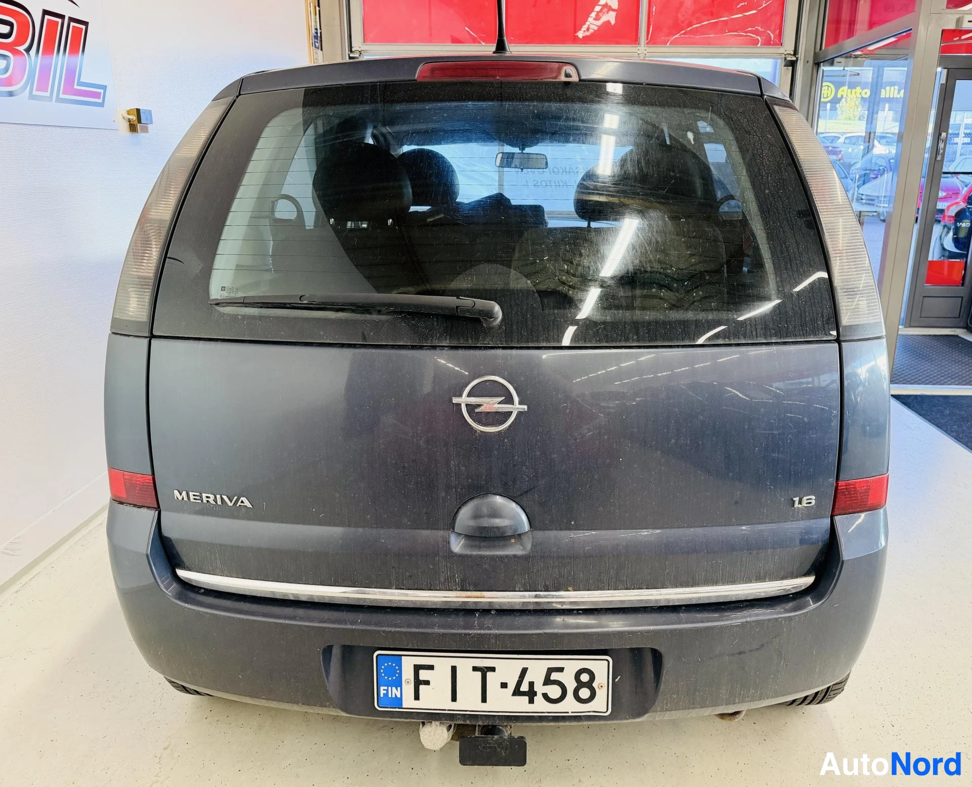Takaa: Opel Merivan takanäkymä sisätiloissa: takaluukussa MERIVA- ja 1.6-merkinnät sekä Opel-logon alla avattava kahva, rekisterikilpi, takaikkunassa pyyhkijä ja pusku