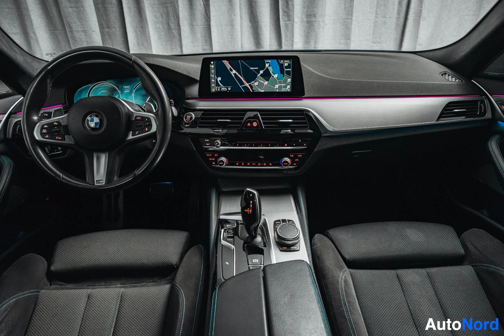 Sivulta: Ohjaamo suoraan eteenpäin kuvattuna: BMW-ohjauspyörä, digitaalinen mittaristo, karttaa näyttävä keskinäyttö kojelaudassa, automaattivaihteen valitsin, iDrive-oh