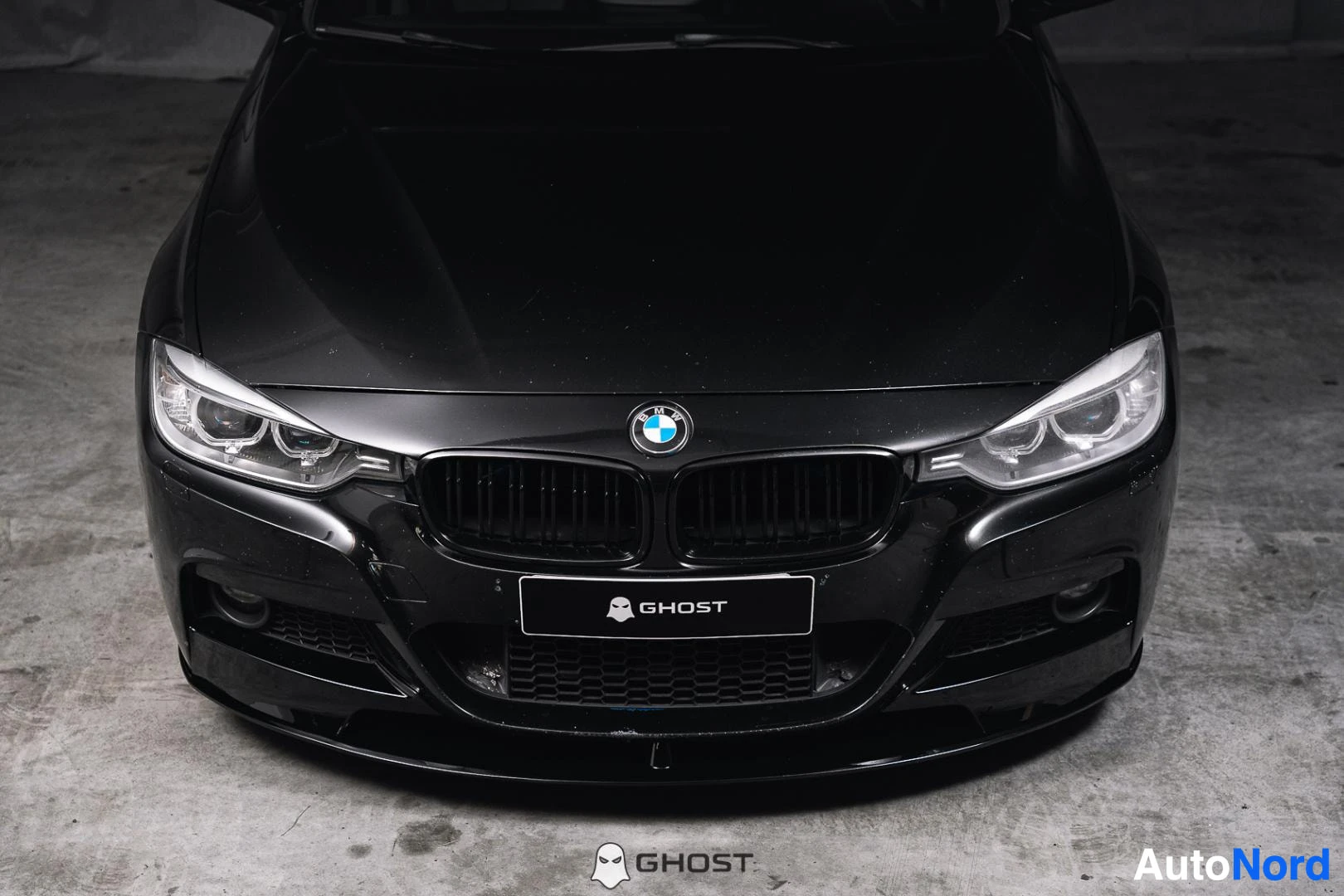Edestä: Yläkulmasta kuvattu mustan auton keula: BMW-etumerkki, ajovalot ja etusäleikkö hallin lattialla.