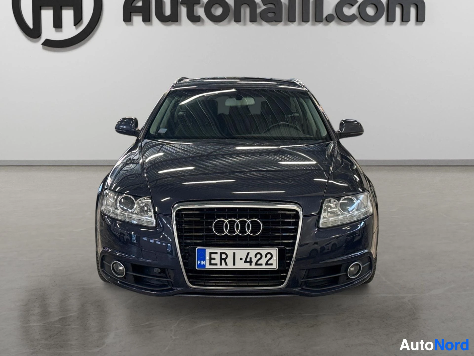 Edestä: Tummansinisen Audi A6:n keulakuva suoraan edestä, Audi-reettikehyksessä neljä renkaan logoa.