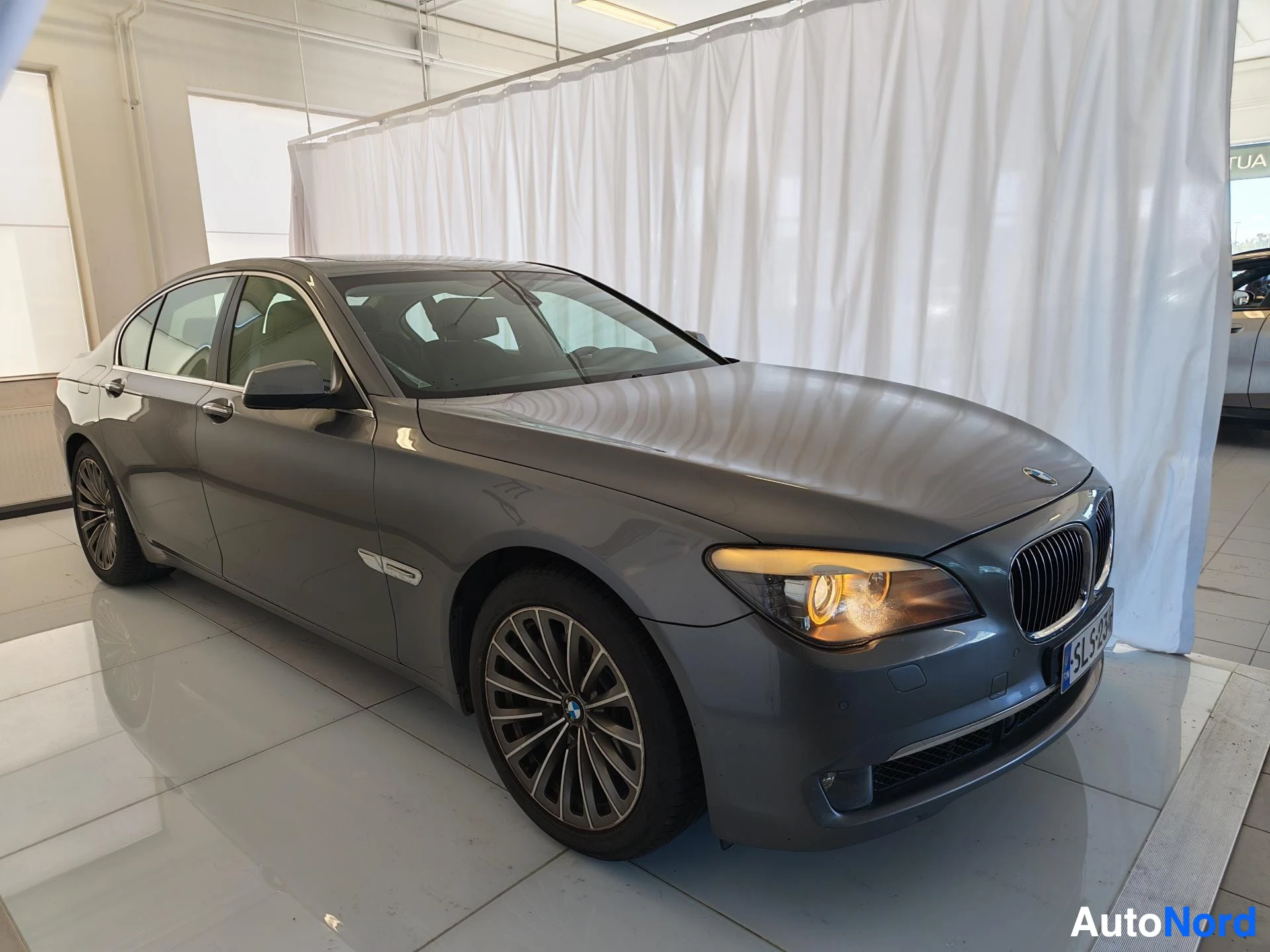 Etuviisto, nokka oikealle: BMW 7-sarjan sedan tummanharmaana kuvattuna kolmeneljännes-etunäkymästä oikealta, myyntitilan valkoisella verholla taustalla.