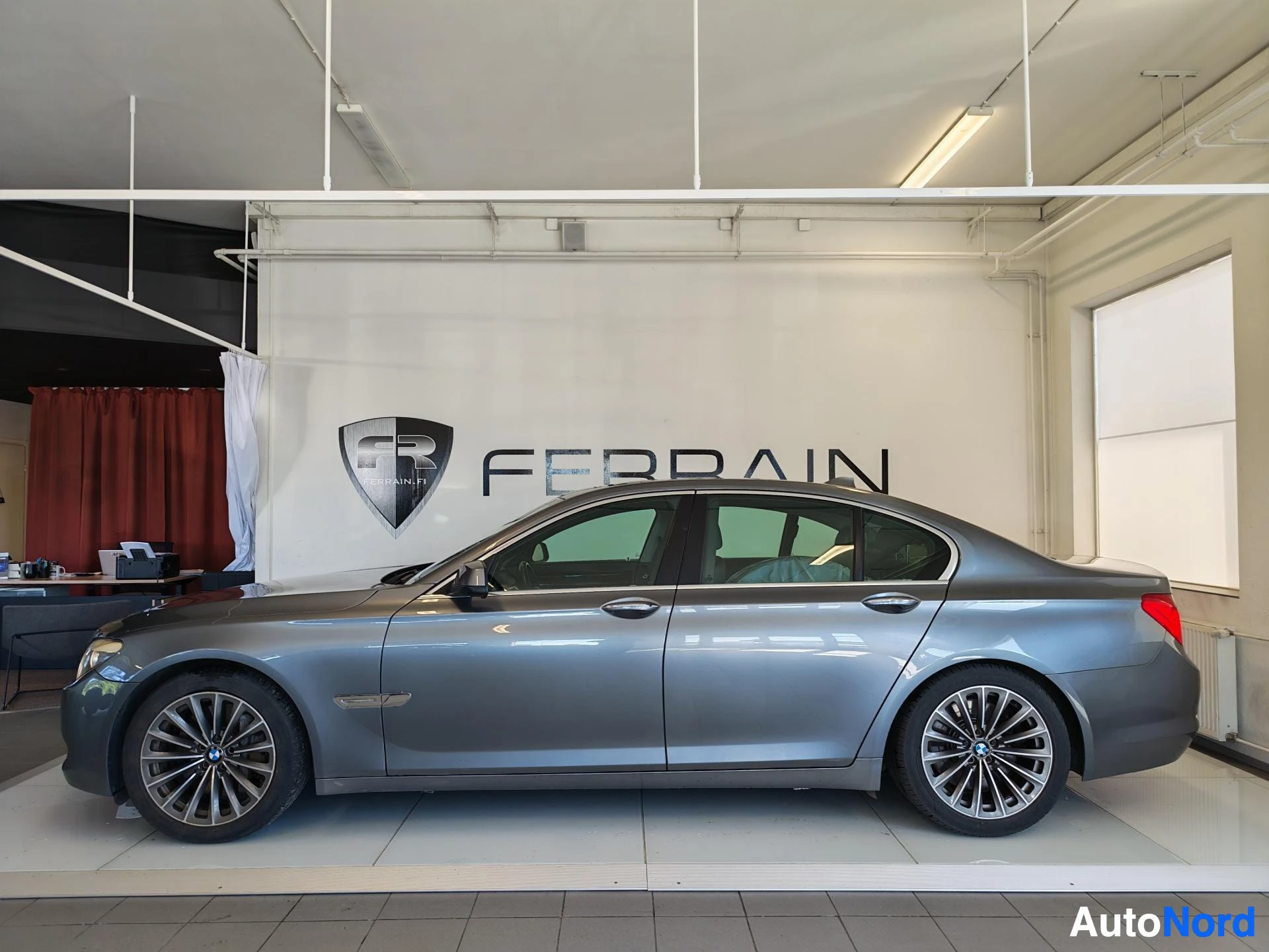Etuviisto, nokka vasemmalle: BMW 7-sarjan sedan sivuprofiili oikealta, FEBRAIN-taustaseinä ja tummanharmaa kori.