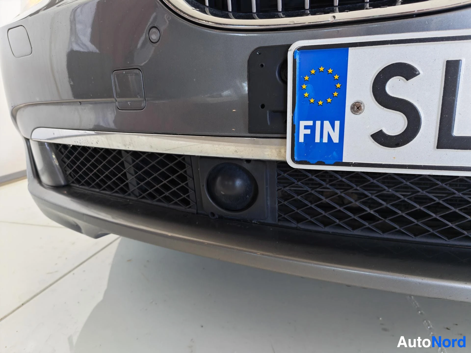 Yksityiskohta: Lähikuva etupuskurin alaosasta, kylttilistalla FIN-tunnus ja EU-tähdet, verkkosäleiköt ja sumuvalaisin.