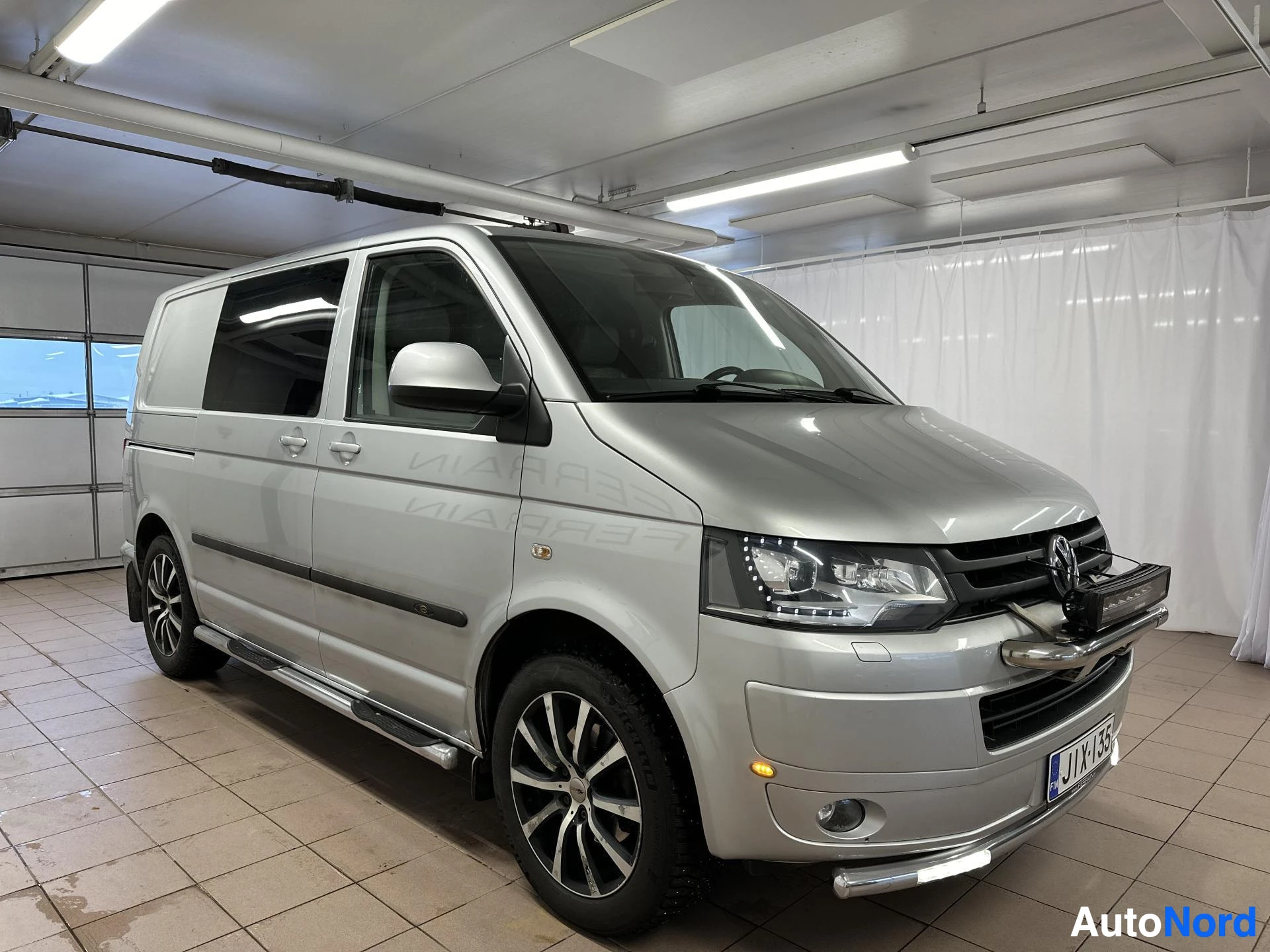 Etuviisto, nokka oikealle: Hopeanvärinen Volkswagen Transporter etuviistosta kuvattuna; etupää ja oikea kylki näkyvissä, etulokasuoja ja kattotelineet ajeleituksessa.