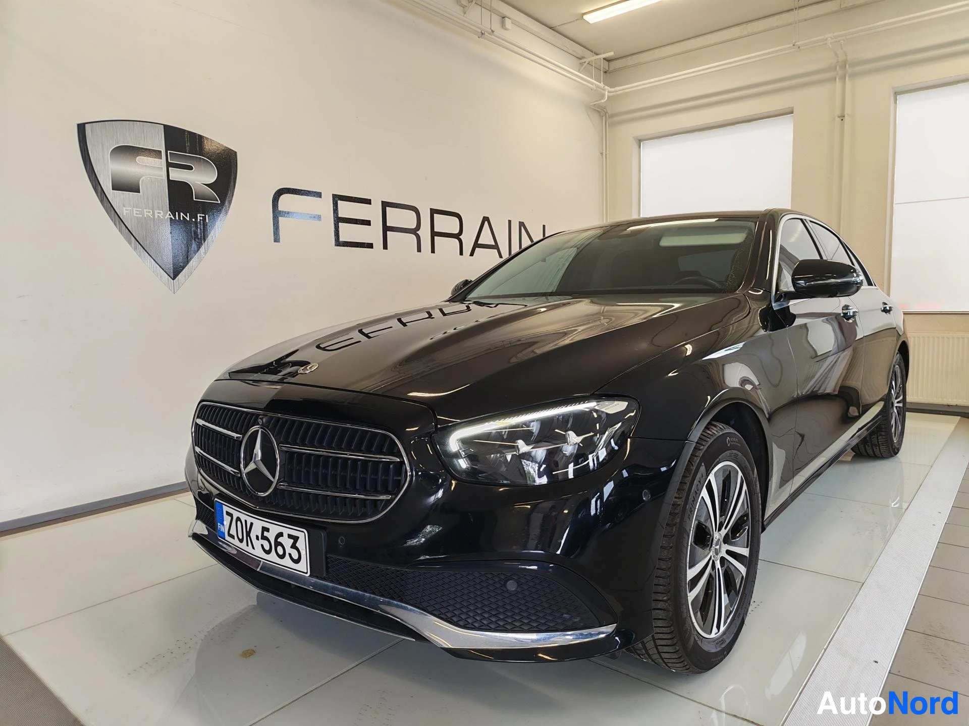 Etuviisto, nokka vasemmalle: Mustan Mercedes-Benz E-sarjan sedanin etupää ja rekisterikilpi myyntitilassa.