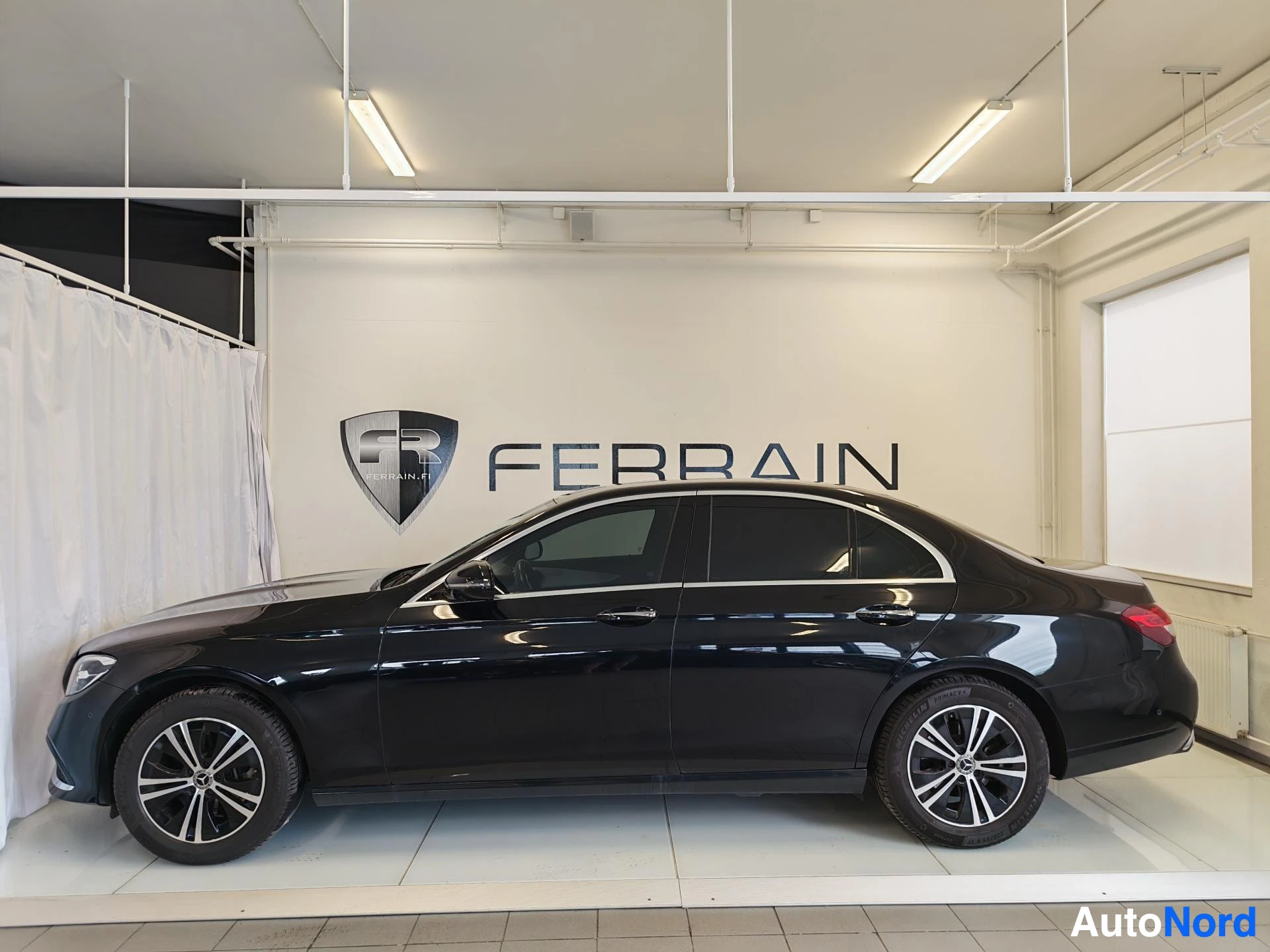 Sivulta: Musta Mercedes-Benz E-sarjan sedan sivulta myyntitilassa, pyörät ja korin sivut näkyvissä.