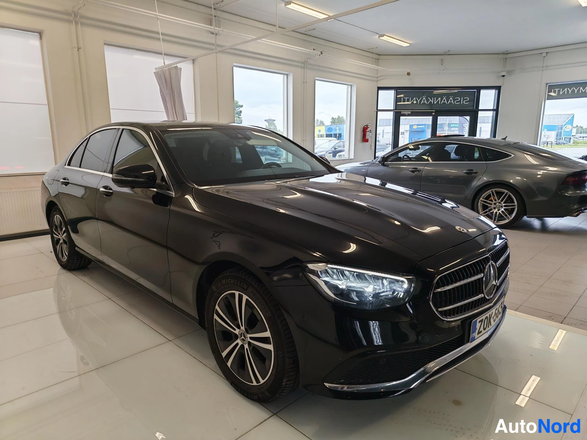 Etuviisto, nokka oikealle: Mustan Mercedes-Benz E -sedanin etukulma autonhallissa, keulakilpi.