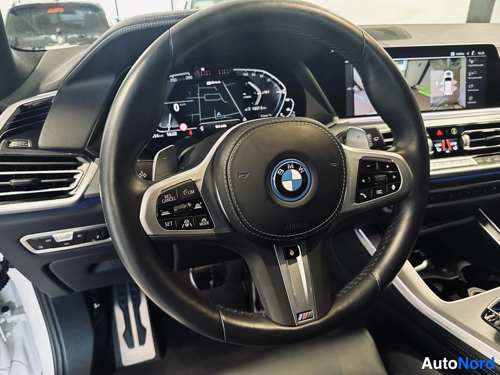 Etuistuimet: Black leather BMW steering wheel with M logo, multifunction controls including cruise buttons and gear shift paddles behind. In the background, a digital dashbo