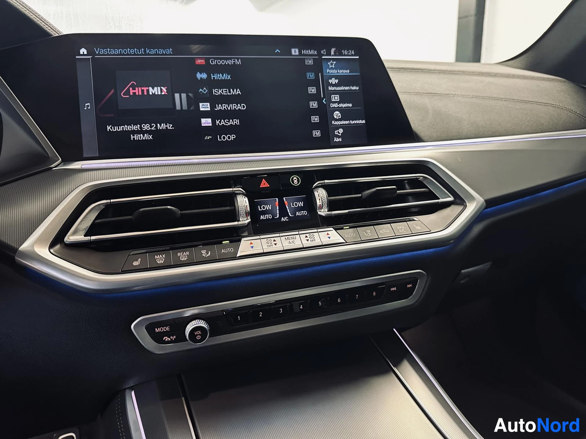 Muu: Auton infotainment-näyttö radiokanavavalikossa, alla ilmastointipaneeli ja keskikonsolin paristot.