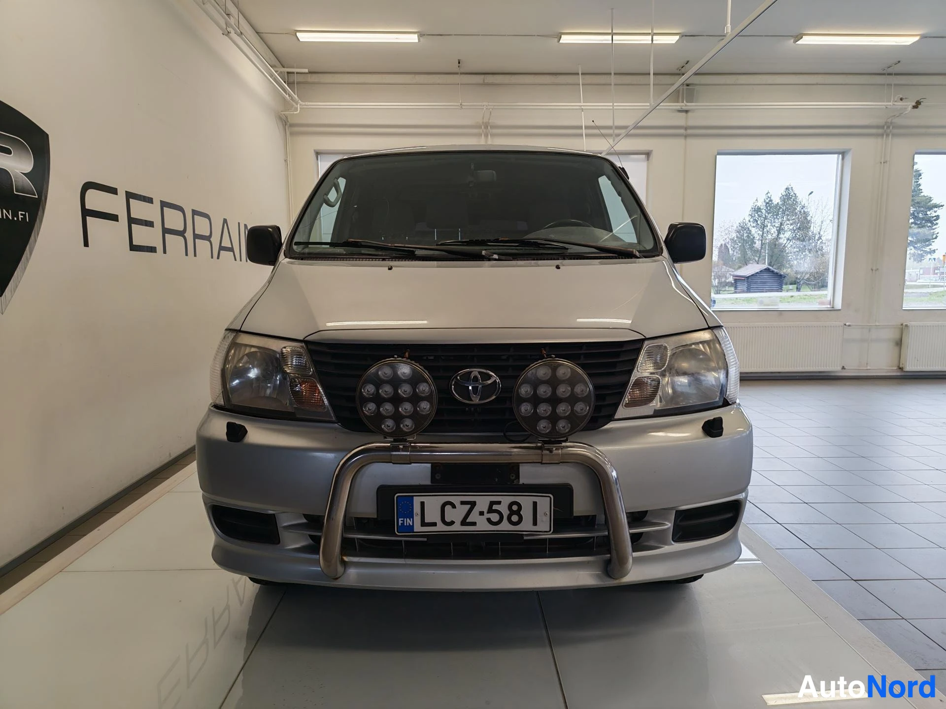 Edestä: Toyota Hiace -pakettiauto suoraan edestä,Toyota-merkki ja lisävalot etusäleikössä, rekisterikilpi.
