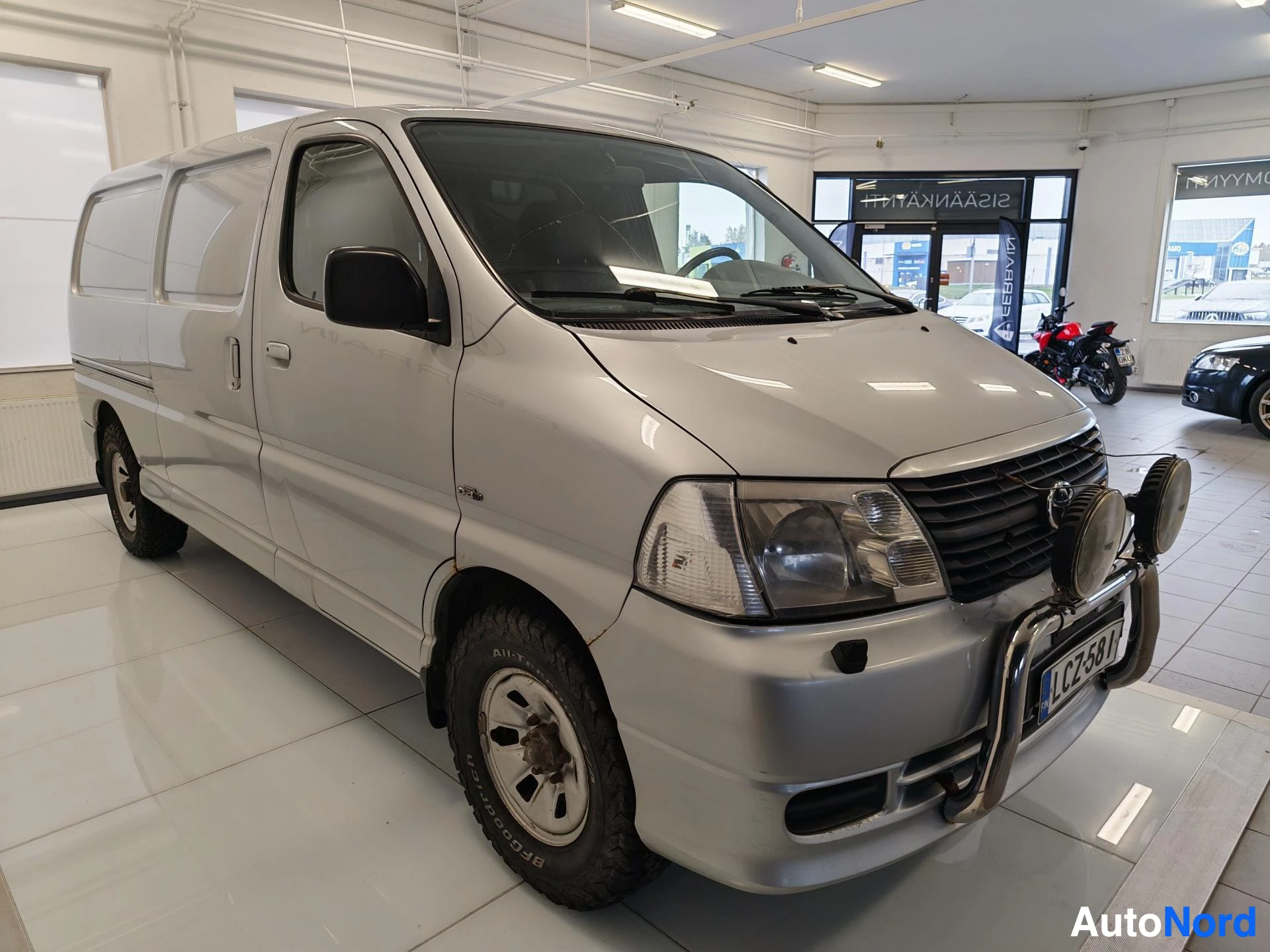 Etuviisto, nokka oikealle: Hopeanhohtoinen Toyota Hiace -pakettiauto etukulmasta oikealta, lisävalot ja rekisterikilpi.