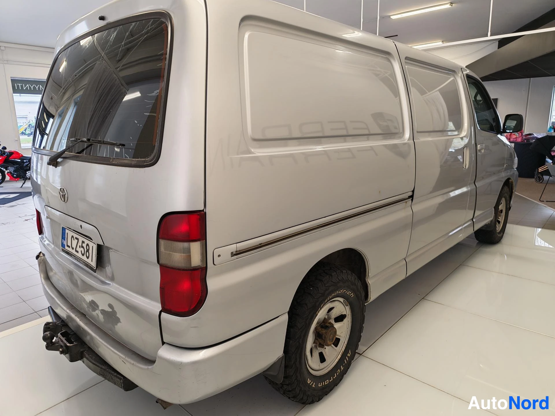 Takaviisto: Toyota Hiace -pakettiauton takakolmasosanäkymä oikealta, hopeinen korilaatikko, rekisterikilpi, takana hinauskestä.