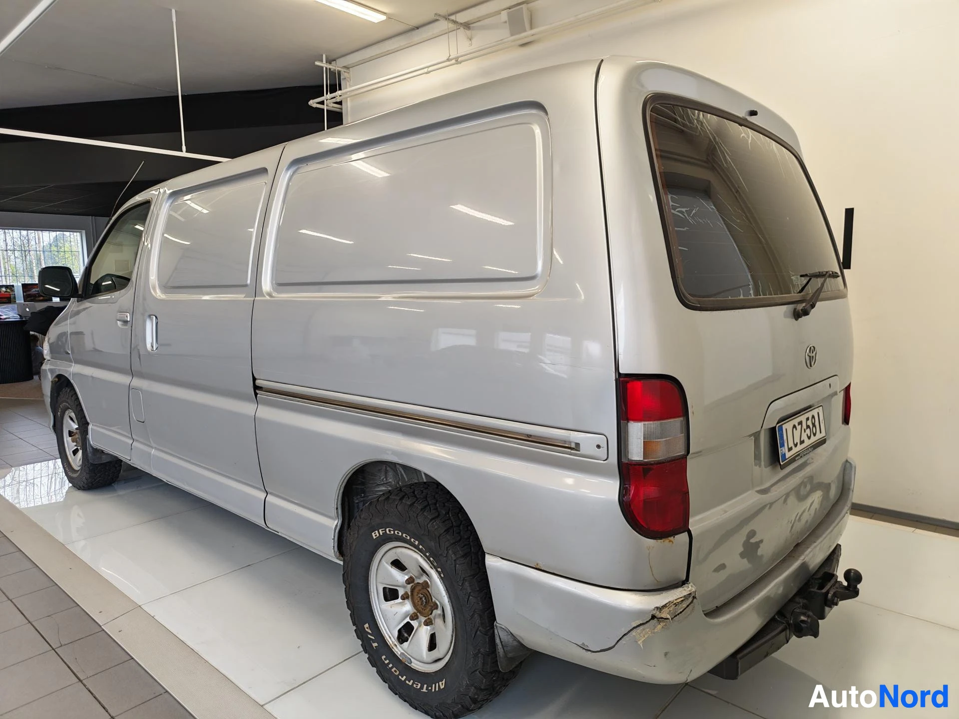 Takaviisto: Toyota Hiace -pakettiauton takakolmasosanäkymä oikealta, hopeinen, BFGoodrich-renkaat, rekisterikilpi, hinauskekki takana.