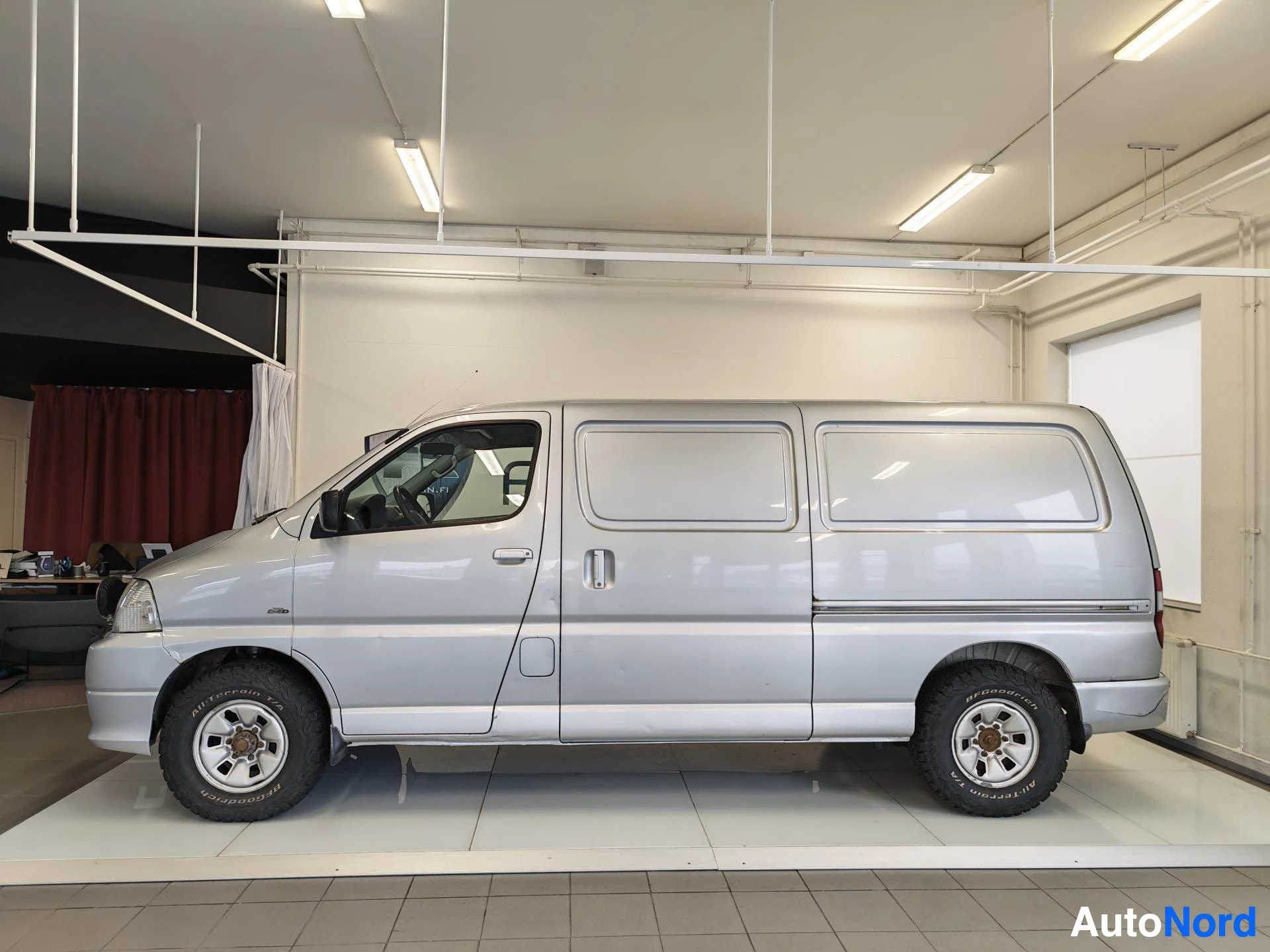 Sivulta: Toyota Hiace -pakettiauton vasen sivuprofiili näytteilleasetustilassa, hopeinen korilaatikko, kolme ovea sivulla.