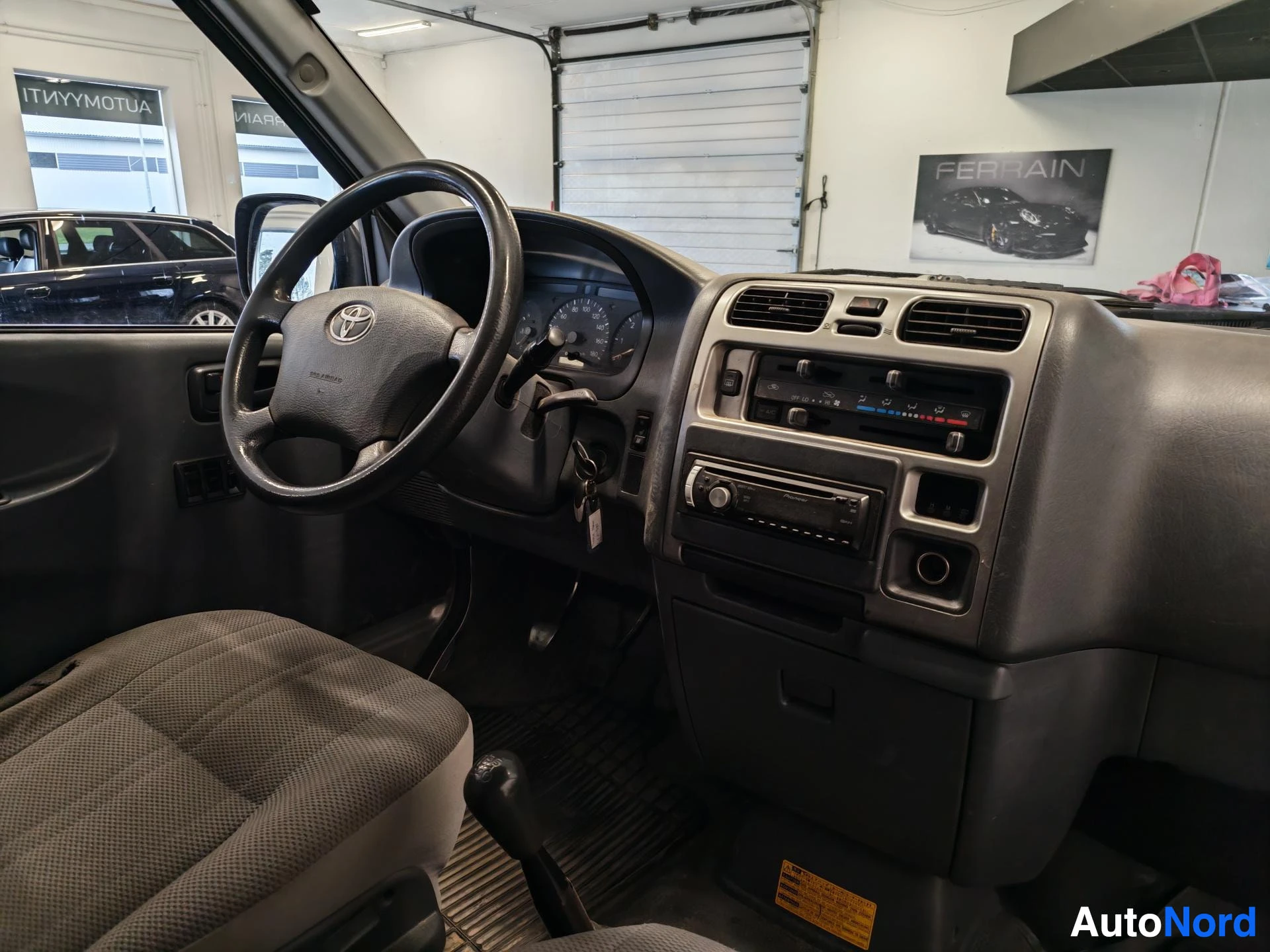 Edestä: Ohjaamo vasemmalta katsottuna: Toyota-ohjauspyörä, mittaristo, radio ja manuaalivaihde.