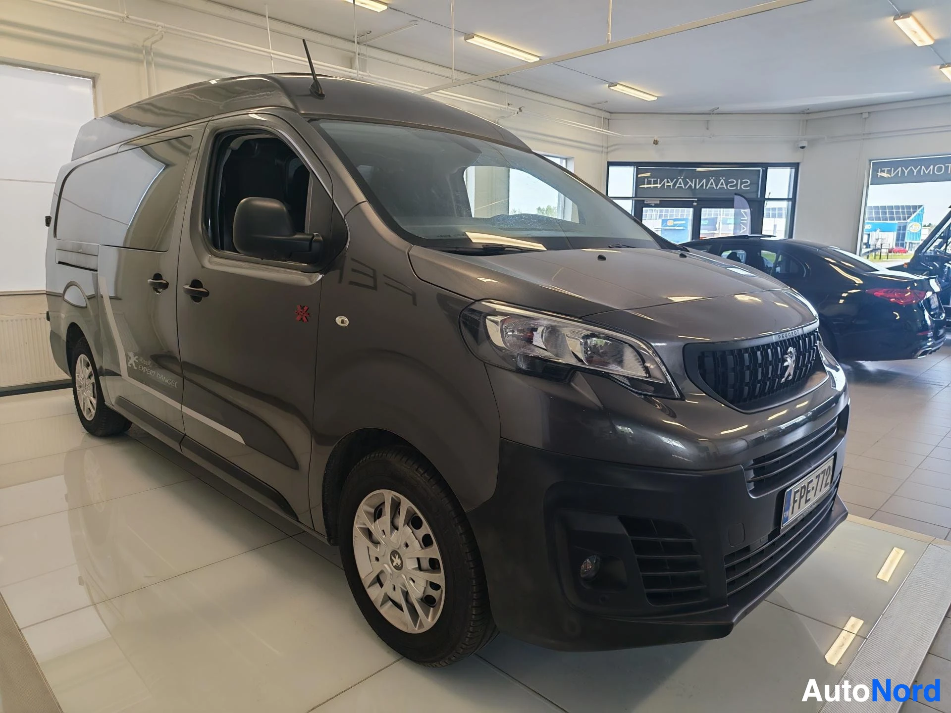 Etuviisto, nokka oikealle: Tummanharmaa Peugeot Expert etuviistossa autosalissa, etusäleikössä Peugeot-leijona ja kilpi.