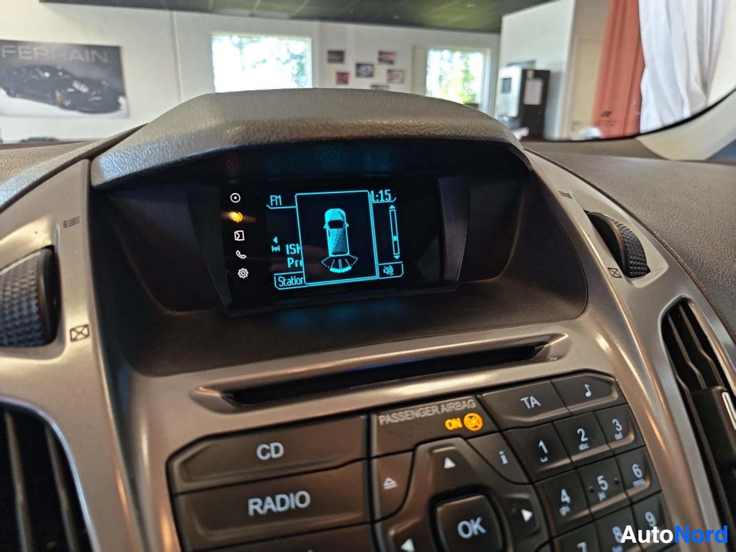 Muu: Auton kojelaudan näyttö ja radio: näytössä FM-viritin ja pysäköintitutkan kuva, alla CD- ja RADIO-napit, numeronäppäimistö sekä PASSANGER AIRBAG -merkintä.