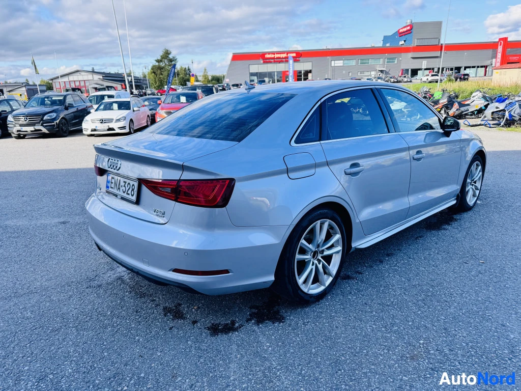 Takaviisto: Hopeinen Audi A3 -sedan takaa vasemmalta sivulta kuvattuna, rekisterikilpi, kauppa­piha taustalla.