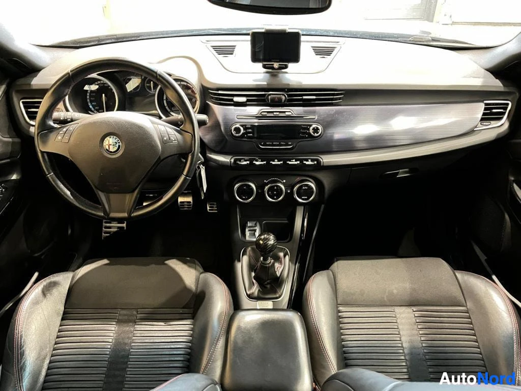 Muu: Alfa Romeon ohjaamo edestä: ratissa Alfa Romeo -logo, manuaalivaihtoheitin, metalliset polkimet, kolme ilmastointisäädintä ja kosketusnäyttö kojelaudan päällä.