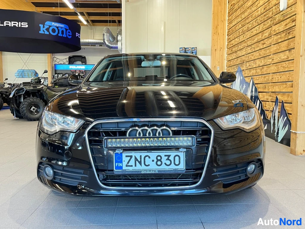 Edestä: Mustan sedanin etunäkymä, rekisterikilpi ja etusäleikkö Audi-renkaalla.