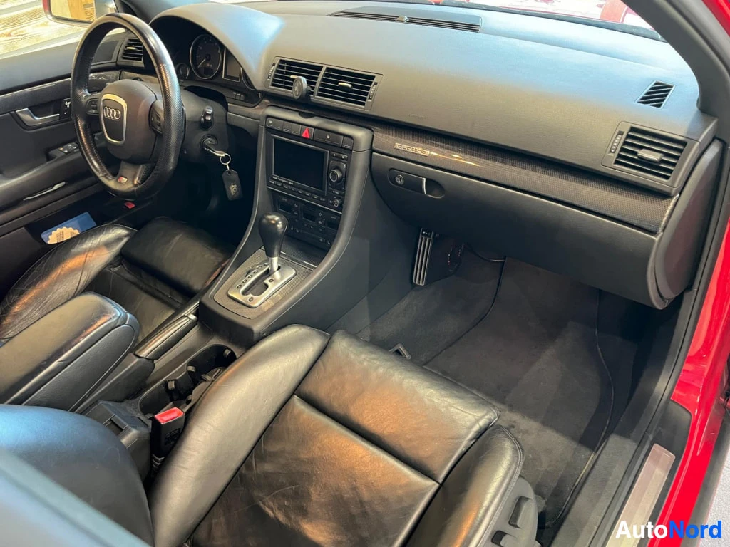 Muu: Kuljettajan puolen näkymä: musta kojelauta, Audi-rattit, automaattivaihteen valitsin ja keskusnäyttö; punainen kori näkyy oviaukossa.