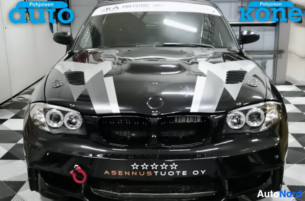 Edestä: Musta BMW:n keulosuora etunäkymä, puskurissa punainen hinauskoukku ja ASENNUSTUOTE OY -kyltti.