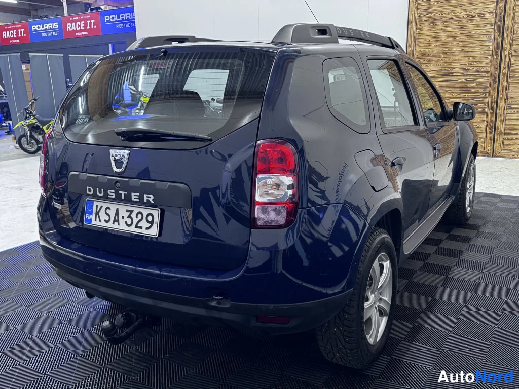 Takaviisto: Back and passenger side of a dark blue Dacia Duster SUV parked on a black grid floor.
