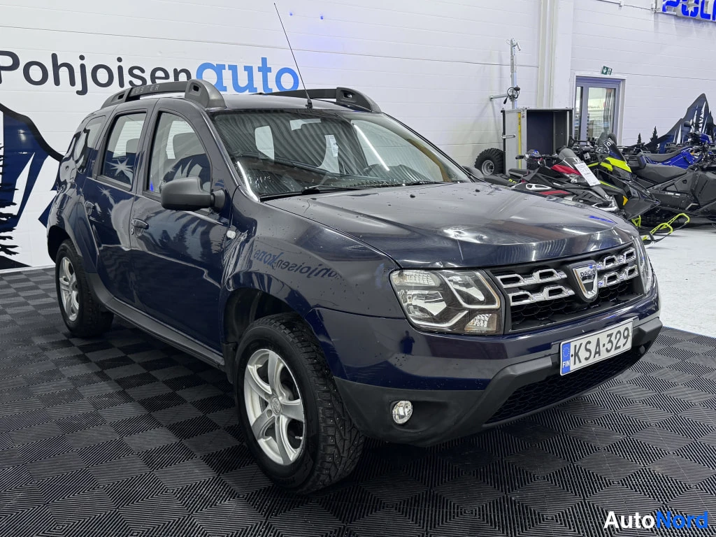 Etuviisto, nokka oikealle: Front and driver's side of a dark blue Dacia Duster with silver roof rails and five-spoke alloy wheels.
