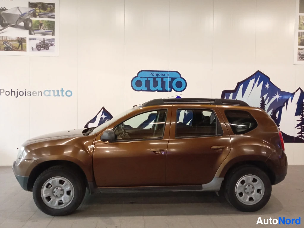 Etuviisto, nokka oikealle: Ruskea Dacia Duster sivusta kulmasta kuvattuna sisätiloissa, kattokaiteet näkyvissä.