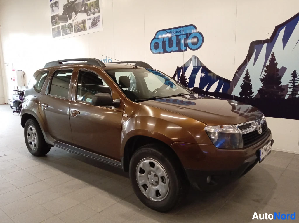 Etuviisto, nokka oikealle: Ruskea Dacia Duster etuviistosta oikealta kuvattuna sisätiloissa, keulakota ja etuvalot näkyvissä.