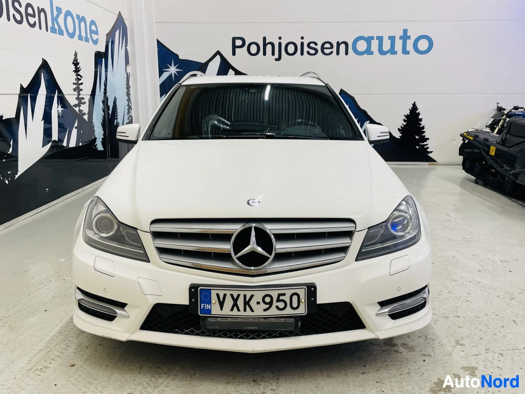Edestä: Mercedes-Benz C-sarjan farmari suoraan edestä, valkoinen kori, ruudukossa Mercedes-tähti, rekisteritunnus.