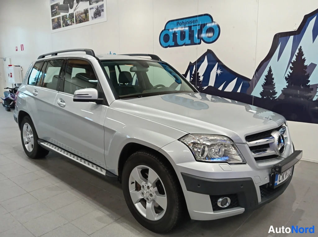 Etuviisto, nokka oikealle: Front three-quarter view of a silver Mercedes-Benz GLK showing its distinctive grille with large emblem, headlights, fog light housings, and front bumper