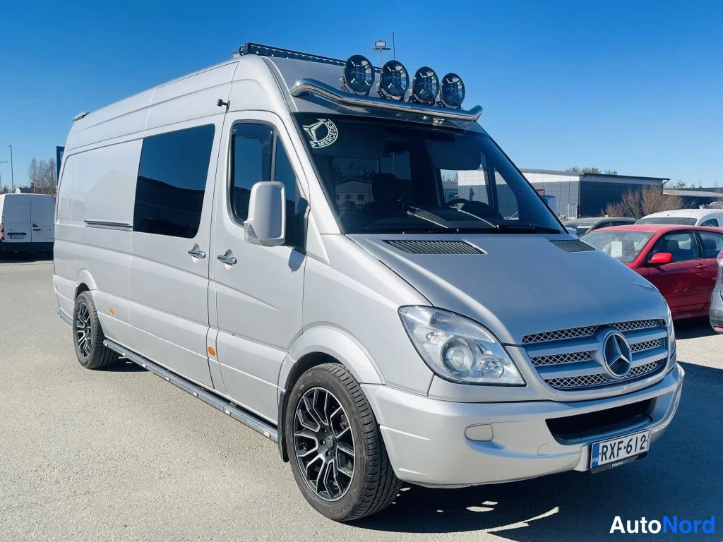 Etuviisto, nokka oikealle: Hopeanvärinen Mercedes-Benz Sprinter -pakettiauto kuvattu kolmivartin etukulmasta; kattoteline ja neljä lisävaloa tuulilasin yläpuolella.