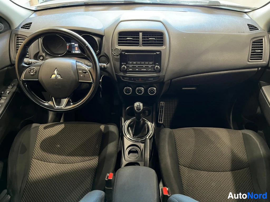 Yksityiskohta: Mittaristö ja tumma kangasverhoiltu sisustus: ohjauspyörä Mitsubishi-merkillä, manuaalivaihteisto ja radio keskikonsolissa.