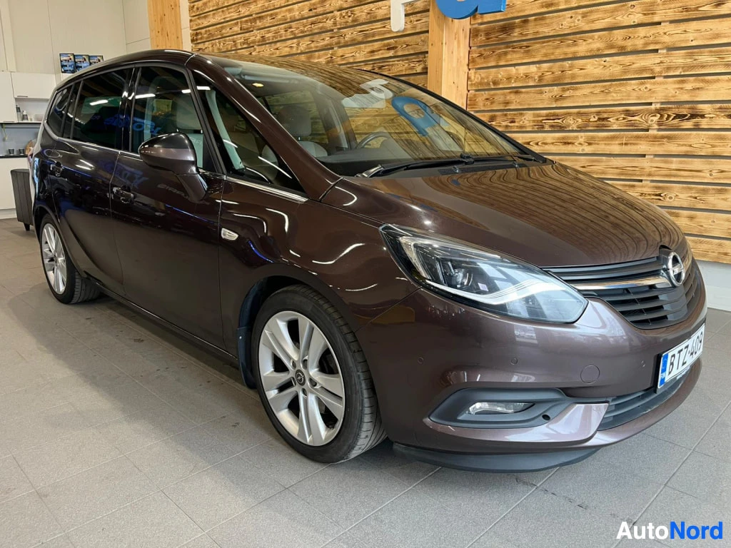Etuviisto, nokka oikealle: Auto edestä oikealta nähtynä: tummanruskean metallivärinen tila-auto, edessä Opel-merkki, sumuvalot ja -kilpi.