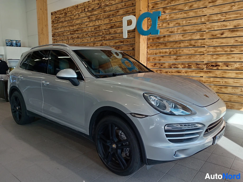 Etuviisto, nokka oikealle: Porsche Cayenne 2011, etukulmakuva oikealta, hopeainen kori, mustat vanteet ja kattokaiteet showroomissa.