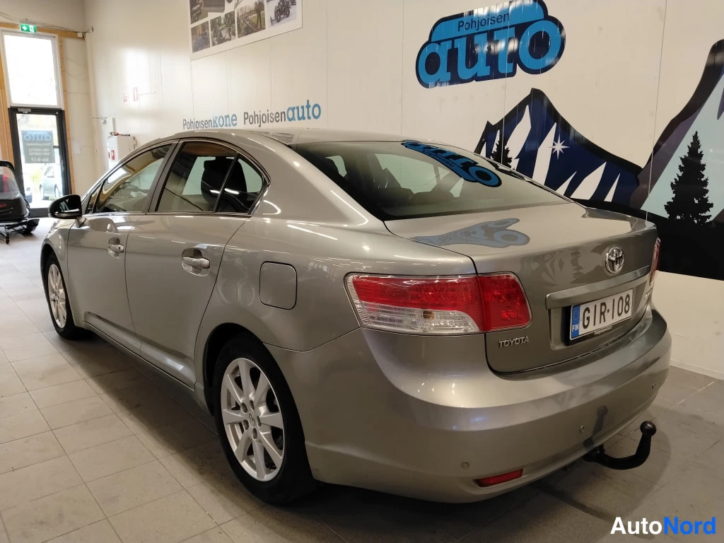 Takaviisto: Toyota Avensis -sedanin taka-vasen kolmannes: punaiset takavalot, TOYOTA-kyltti ja rekisterikilpi tavaratilan kannessa, nostokoulu takapuskurissa.
