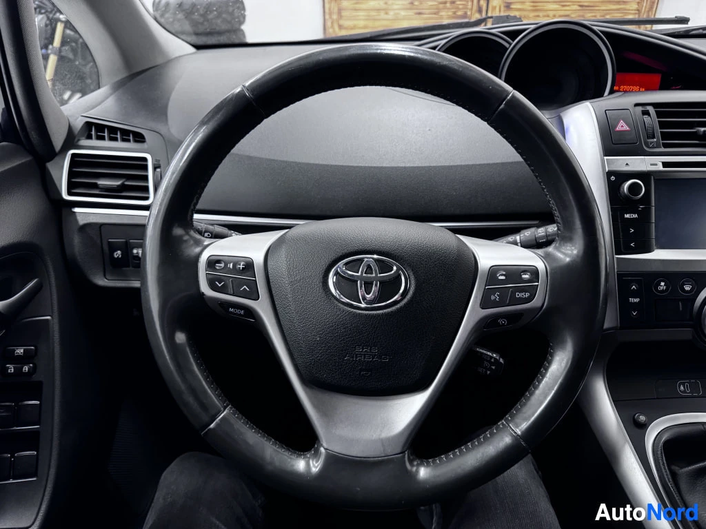 Ohjaamo: Ohjauspää kuva lähikuva. Toyota-logo keskellä. Ohjauspyörässä on painikkeet sekä äänentoistolle että puhelulle. Taustalla mittaristo on hämäränä näkyvissä.