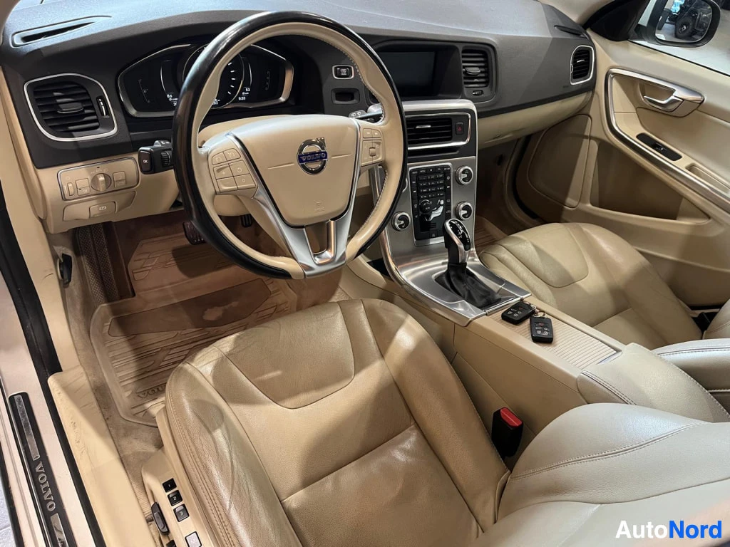 Etuistuimet: Volvo V60:n kuljettajan puolen sisätila: beige näyttö, Volvo-ohjauspyörä, automaattivaihde ja keskosella avaimet.