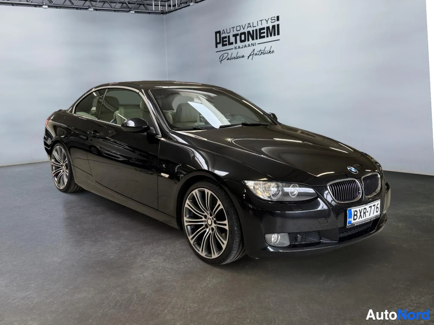 Etuviisto, nokka oikealle: Musta BMW-avomalli kulmassa etuosa oikealle kuvattuna sisätiloissa, rekisteritunnuksella.