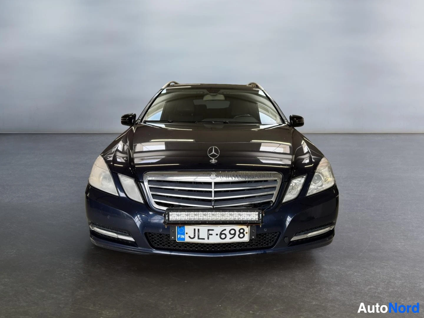 Edestä: Mercedes-Benz E-sarjan farmari suoraan edestä, tummansininen koripinta, rekisterikilpi ja etupuskassa kiinnitetty lisävalopalkki.