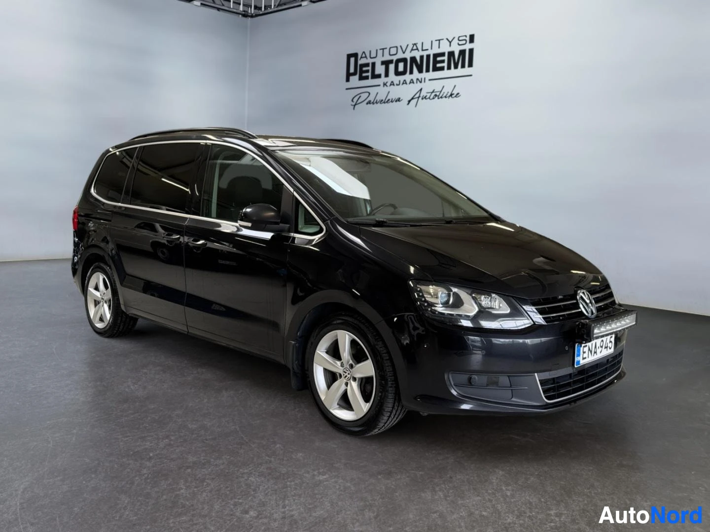 Etuviisto, nokka oikealle: Musta Volkswagen Sharan -tilunen sisätiloissa, etukolmannesvasen kulma, kevytmetallivanteet ja kattokaiteet näkyvissä.