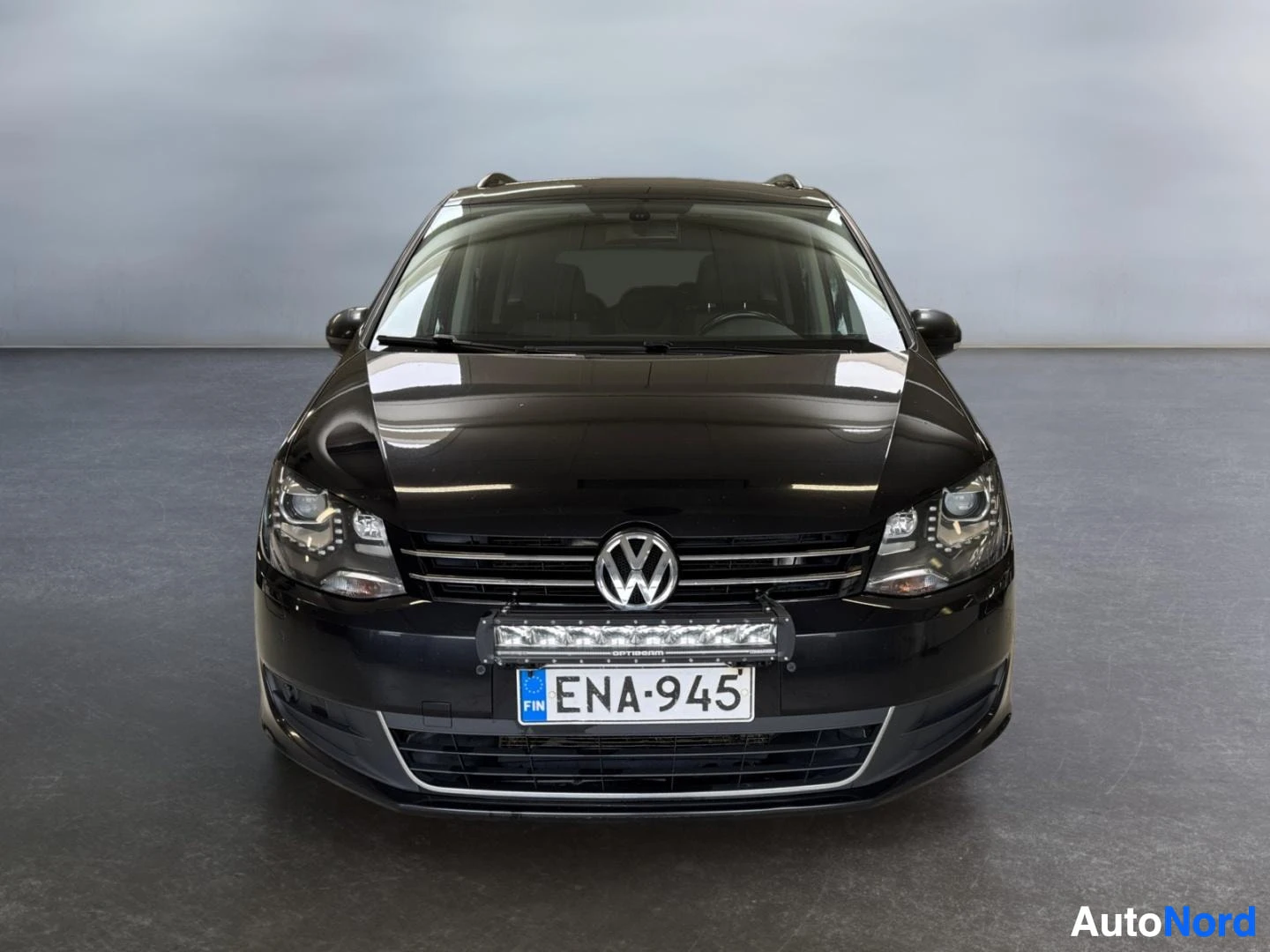 Edestä: Mustan tilavaunun suora etukuva; VW-merkki, lisävalokisko etupuskurissa ja rekisteritunnus.