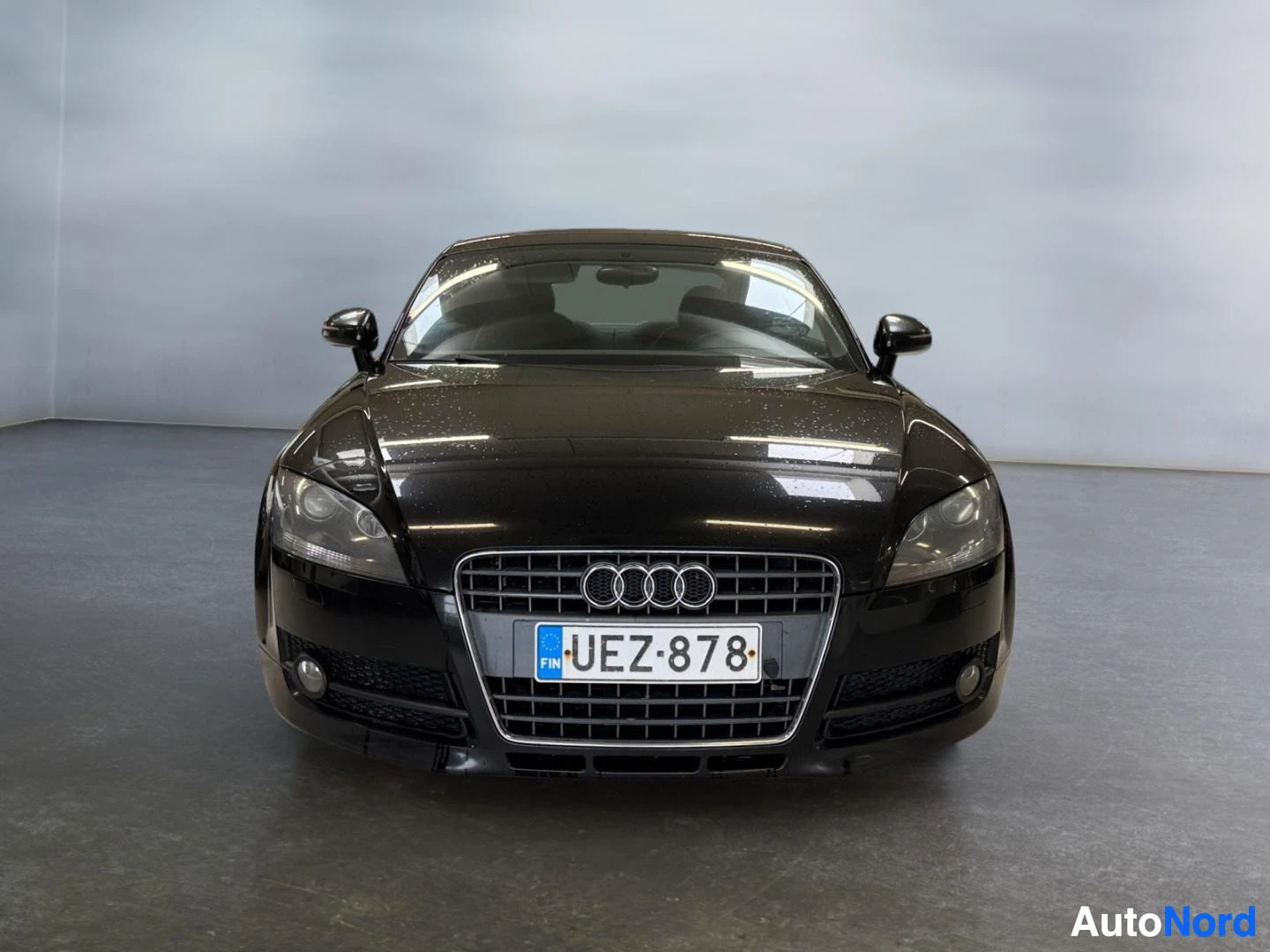 Edestä: Mustan Audi TT Coupén suora etukuva, Audi-tunnuksinen etusäleikkö ja rekisterikilpi näkyvät sisätiloissa.
